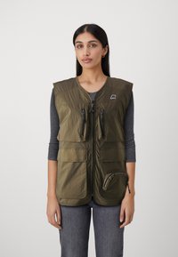 Olīvu zaļš veste, kas izgatavota no viegla materiāla, ar rāvējslēdzēja aizdari, divām priekšējām kabatām un logotipa detaļu augšējā kreisajā pusē.