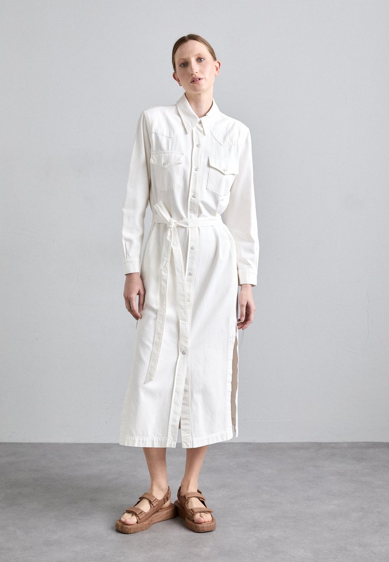 Balta mygtukinė suknelė su diržu, ilgomis rankovėmis, dviem krūtinės kišenėmis, šoniniais įpjovimais ir rudais platforminiais sandalais. Lygus audinio paviršius.