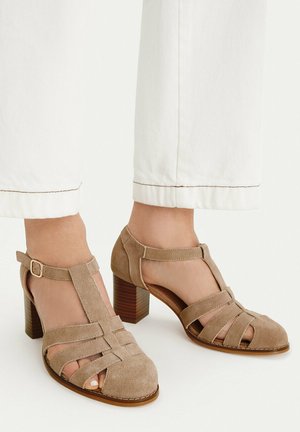 Beige Veloursleder T-Riemchen Sandalen mit Blockabsatz, getragen zu weißen, gekürzten Hosen, mit mehreren vorderen Riemen und seitlicher Schnallenverschluss.