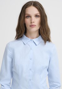 Svijetloplava košulja s dugim rukavima i dugmetima koja ima klasičan ovratnik, glatku teksturu i sedam usklađenih dugmadi duž prednje strane.