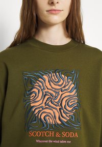 oliwkowy sweter z teksturowanym wzorem w kolorze pomarańczowym i niebieskim, zawierający napis "SCOTCH & SODA" oraz hasło poniżej wzoru.