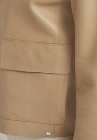 Beige Lederjacke mit einer großen Tasche an der Seite, sichtbaren Nähten und einem kleinen metallischen Label am Saum. Glatte Textur.