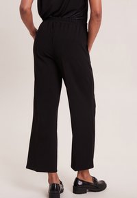 Zwarte wijde broek met een gladde textuur, elastische tailleband en zijnaad. Draagt met glanzende zwarte schoenen met een lage blokhak.