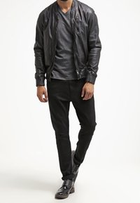 Uomo che indossa una giacca di pelle nera, una maglietta grigia scura, pantaloni neri e scarpe eleganti nere che cammina su uno sfondo bianco.