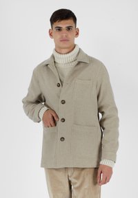 Giacca di lana beige con colletto, chiusura a bottoni e due tasche frontali. Indossata sopra un dolcevita in maglia color crema. Tessuto strutturato, vestibilità casual.