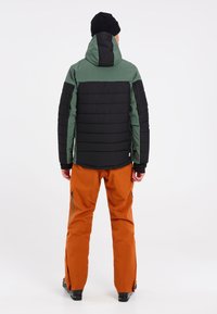 Gewatteerde jas met een zwart ondergedeelte en een groen bovengedeelte, gecombineerd met oranje skibroek met ritszakken en verstelbare manchetten.
