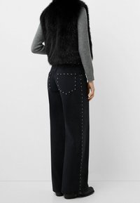 Mustat leveälahkeiset farkut, joissa on hopeiset niittidetaljiet taskujen ja sauma sivuilla; yhdistettynä lyhyeen mustaan tekoturkisliiviin ja harmaan väriseen neuleeseen.