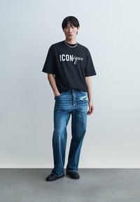 Mladý muž v čiernom tričku s nápisom "ICONique", modrých roztrhaných džínsach, čiernych topánkach a s náhrdelníkom z korálok na jednoduchom svetlom pozadí.