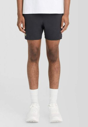 Persoon die zwarte korte broek, witte sokken en witte sneakers draagt, staand tegen een effen lichte achtergrond.