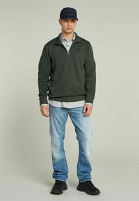 Sudadera verde oscuro con un cuello de media cremallera, superpuesta a una camiseta de rayas gris claro, combinada con vaqueros azules claros y zapatillas negras.