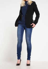 Černý blazer přes námořnický top, spárovaný s úzkými modrými džínami a černými vysokými podpatky. Blazer má jeden knoflík a otevřený přední díl.