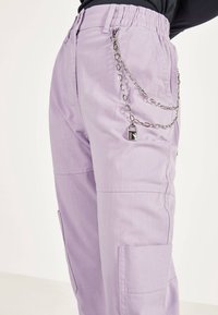 Lilawe spodnie cargo z elastycznym pasem, bocznymi kieszeniami oraz dekoracyjnym srebrnym łańcuszkiem z kłódką zawieszonym na pętli paska.