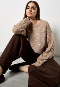 Maglione lavorato a maglia color marrone chiaro con una leggera lucentezza, polsini a costine e vestibilità morbida, abbinato a pantaloni larghi marrone scuro. Sfondo in cemento.