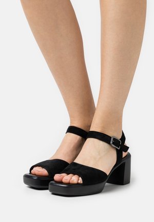 Sandales en daim noir à talon carré avec un design à bout ouvert, bride de cheville avec boucle en métal et une silhouette moderne et lisse.
