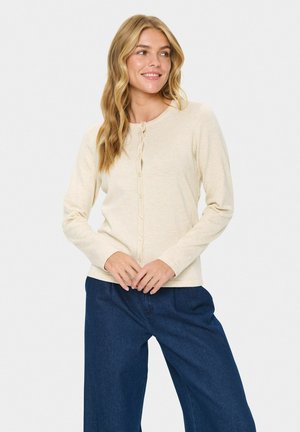 Beige, langærmet cardigan med rund halsudskæring, forreste knaplukning og ribbede manchetter, parret med vide mørkeblå bukser.