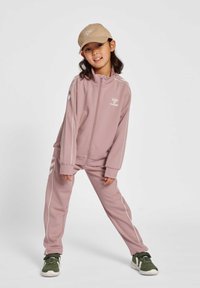 Ljusrosa träningsoverall med vita detaljer och logotyp, bestående av en zip-up jacka och raka byxor. Matchas med en beige keps och gröna sneakers.