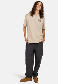 Beige cotton t-shirt with short sleeves and a printed logo, paired with dark cargo pants featuring pockets and a relaxed fit. Brown shoes included.