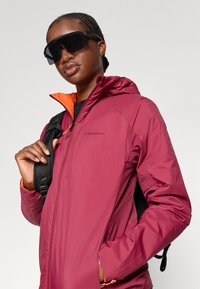Maroon vedenpitävä takki, jossa on oranssi sisävuori, huppu ja säädettävät hihnat. Musta reppuolkalaukun hihna näkyvissä, malli käyttää tummia aurinkolaseja.