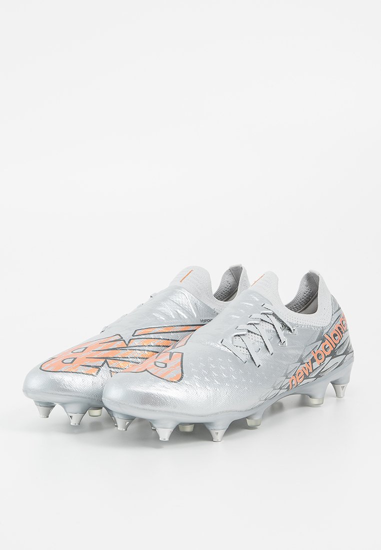 Crampons de soccer argentés avec une tige synthétique texturée, accents orange, fermeture à lacets et crampons moulés pour une adhérence sur les surfaces en gazon.