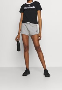 Musta lyhythihainen T-paita, jossa on valkoinen "Reebok"-logo, yhdistettynä harmaisiin shortseihin ja mustiin urheilukenkiin, pidellen mustaa vesipulloa.