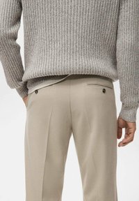 Pull gris clair côtelé associé à un pantalon beige sur mesure avec deux poches arrière et un détail bouton. Texture du tissu lisse visible.