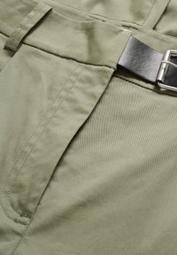 Gros plan sur un pantalon kaki avec poche avant visible, passants pour ceinture, et une ceinture en cuir noire avec boucle en métal.
