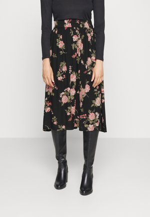 Kobieta ubrana w czarną kwiatową spódnicę midi, czarną bluzkę z długim rękawem oraz czarne skórzane kozaki za kolano, stojąca na jednolitym tle.