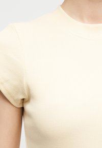 Κοντινό πλάνο ενός ατόμου που φοράει μια ανοιχτό μπεζ ριγωτή κοντομάνικη μπλούζα με στρογγυλή λαιμόκοψη, σε ουδέτερο φόντο.