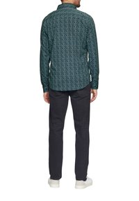 Groen gepatteerd overhemd met lange mouwen en een button-down kraag, gecombineerd met donkere jeans en witte sneakers. De achterkant toont complexe ontwerpdetails.