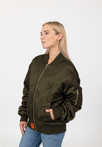 Giacca bomber invernale trapuntata verde oliva con zip frontale, polsini a costine e tasche laterali. Presenta un piccolo etichetta arancione del brand sul davanti.