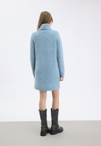 Svetlo modré, štruktúrované šaty sveter s vysokým golierom, dlhými rukávmi a rovnou siluetou. Spárované s čiernymi čižmami po kolená.