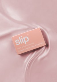 Rosa boks merket "glide" med produktdetaljer, hviler på en glatt, lys rosa silkeoverflate. Teksten nevner anti-aldring og søvnfordeler.