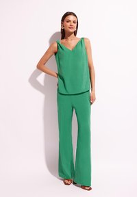 Zielony bezrękawnik z dekoltem w serek i związanymi ramiączkami zestawiony z szerokimi spodniami, wszystko w gładkim, lekki materiale. Brązowe sandały dopełniają całość.
