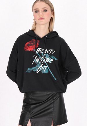 Młoda kobieta nosząca czarną bluzę z kapturem z czerwonym różą i napisem „Beauty Inside Out”, w połączeniu z czarną skórzaną spódnicą z przednimi rozcięciami.