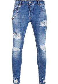 Kék farmer szűkített nadrág, elhasználódott foltokkal, rojtos szegélyekkel és ezüst lánc díszítéssel a bal zseben. Klasszikus öt zsebes kialakítás.