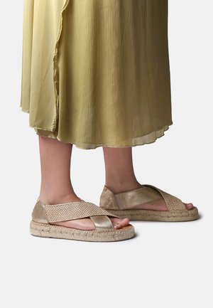 Voeten met gouden espadrille sandalen met gevlochten bandjes, gecombineerd met een vloeiende lichtgroene geplooide rok.