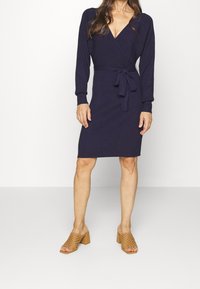 Abito avvolgente blu marino con maniche lunghe, una cintura da annodare in vita e una texture a maglia liscia. Abbinato a scarpe con tacco a blocco aperte beige.