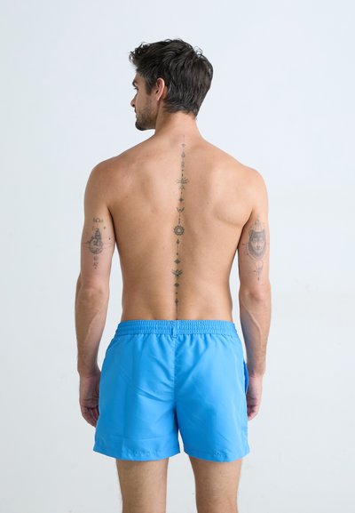 Shorts de bain bleus en tissu léger, dotés d'une taille élastique et de poches latérales. Tatouages visibles sur le corps le long de la colonne vertébrale et des bras.