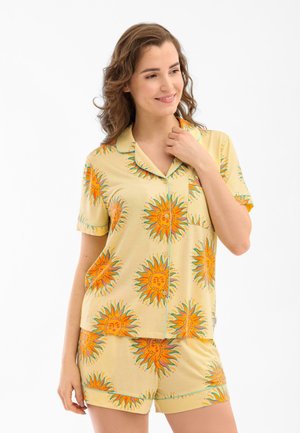 Woman wearing yellow short-sleeve pajama set with orange sun patterns, standing against a white background, smiling and touching collar.