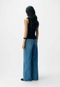 Schwarzes ärmelloses Oberteil kombiniert mit weiten blauen Jeans, die eine hohe Taille und Fading zeigen. Schwarze Schuhe und ein Armband runden den Look ab.