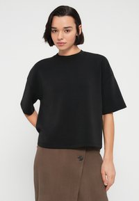 Μαύρο κοντομάνικο μπλουζάκι με φαρδιά σιλουέτα και στρογγυλή λαιμόκοψη, κατασκευασμένο από απαλό ύφασμα. Συνδυασμένο με καφέ φούστα που έχει λεπτομέρεια με κουμπί.