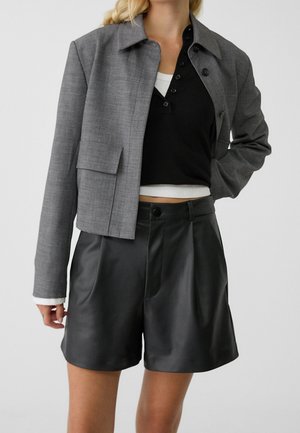 Kobieta w szarej krótkiej kurtce na warstwowych czarno-białych topach, w zestawieniu z czarnymi skórzanymi szortami z wysokim stanem, stoi z jedną ręką na biodrze.