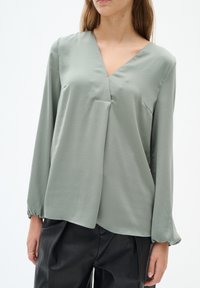Blusa verde chiaro con scollo a V, maniche lunghe e vestibilità ampia. Realizzata in un tessuto morbido, presenta leggeri drappeggi e polsini elasticizzati.