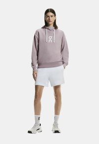 Sudadera con capucha lila con logo blanco y cordones, combinada con pantalones cortos blancos y zapatillas. La tela parece suave y ligeramente texturizada.