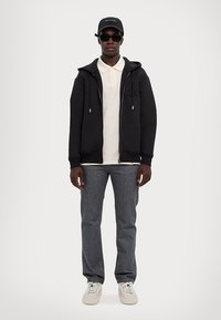 Felpa nera con zip e cappuccio, polo bianca sotto, jeans grigi, sneakers bianche e cappellino nero con occhiali da sole.