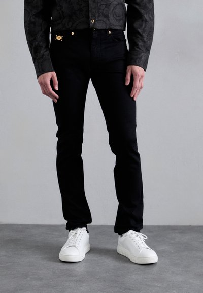 Zwarte spijkerbroek met een slim fit, gouden hardwareaccenten, gecombineerd met witte sneakers. De jas heeft een gestructureerd zwart patroon.