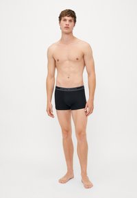 Schwarze Boxershorts aus elastischem Material mit einem markierten Bund. Das Modell steht barfuß und zeigt ein tailliertes Design und eine glatte Textur.
