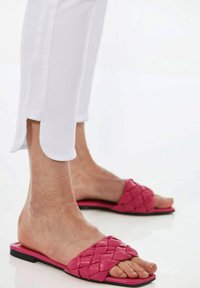 Rosafarbene, geflochtene Rutsch-Sandalen mit einer flachen schwarzen Sohle. Getragen mit weißen, maßgeschneiderten Hosen mit geschlitzten Säumen am Knöchel.