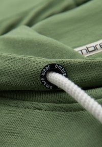 Tela de algodón verde con un acabado texturizado, que presenta un ojete negro con un cordón blanco y una etiqueta con la impresión "ombre wear".