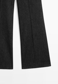Zwarte denim jeans met brede uitlopende pijpen, getoond vanaf de onderkant tegen een witte achtergrond.